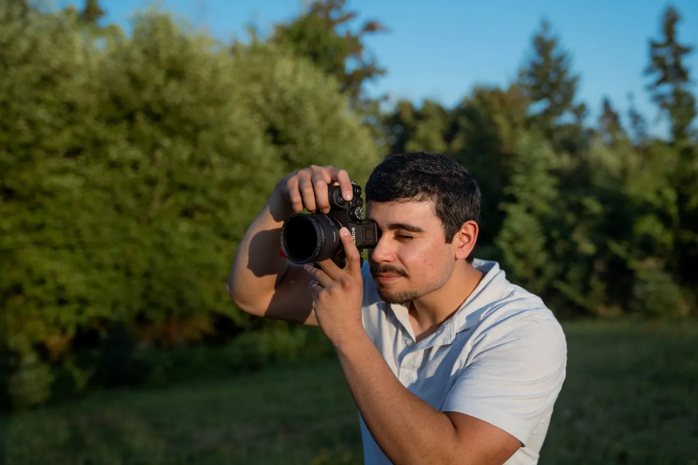 DJ Riley looking through the viewfinder of his Sony camera in a sunlit meadow